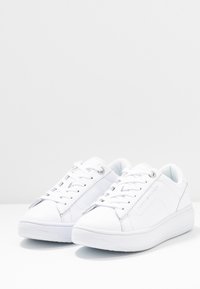 Białe sneakersy z zapięciem na sznurówki, wykonane z materiału syntetycznego, o gładkiej fakturze, zaokrąglonym nosku i subtelnym logo z boku. Płaska gumowa podeszwa.
