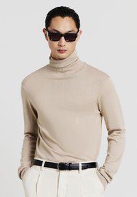 Beige turtleneck sweater with long sleeves, made from a smooth fabric. Worn with black belt and white trousers, complemented by dark sunglasses.