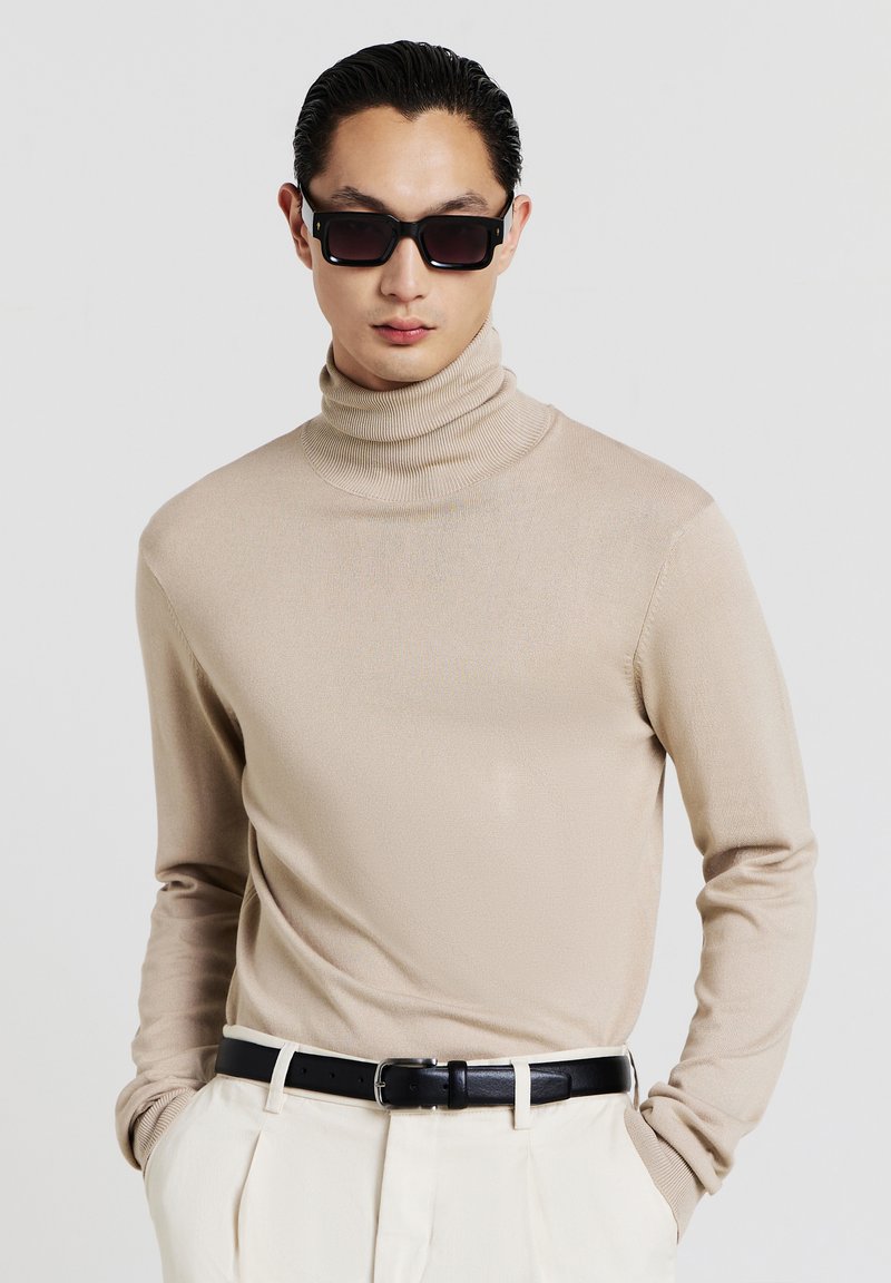 Beige turtleneck sweater with long sleeves, made from a smooth fabric. Worn with black belt and white trousers, complemented by dark sunglasses.