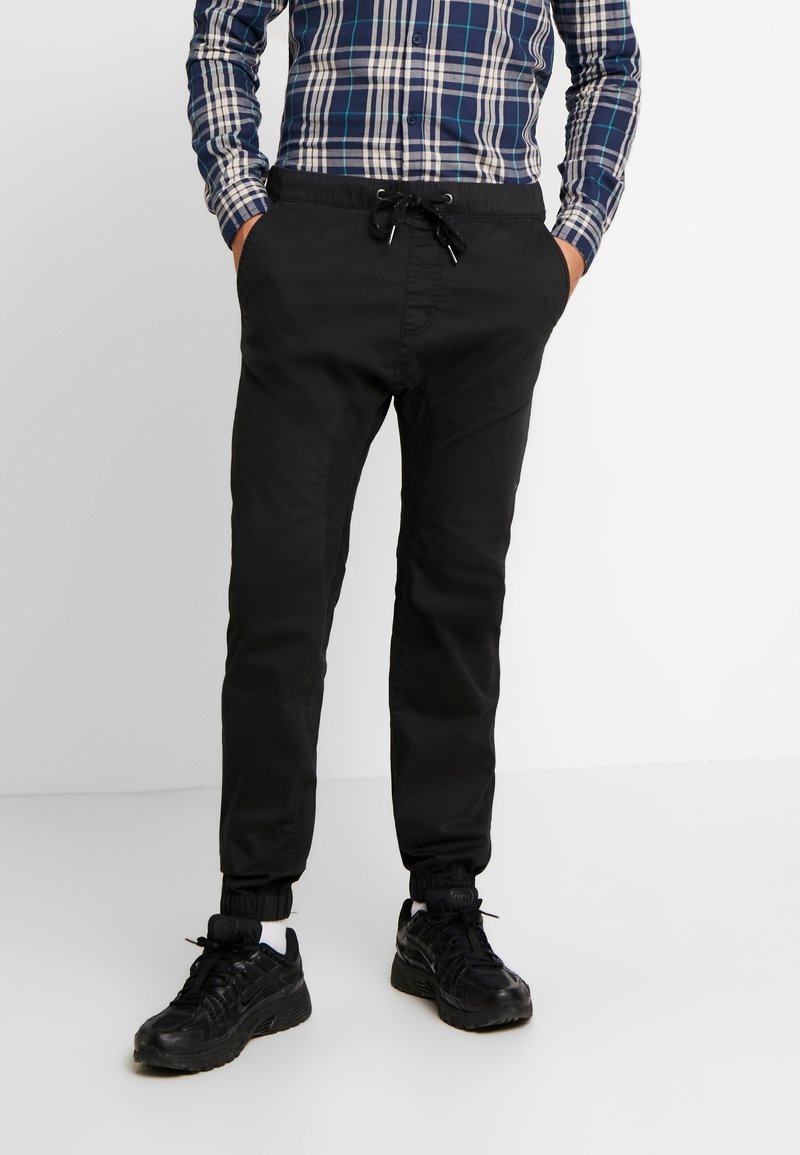 Homme portant un pantalon de jogging noir à cordon, des baskets noires, des chaussettes blanches et une chemise à manches longues à carreaux bleu et blanc, les mains dans les poches.