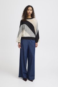 Pull en gros format, multicolore, avec des motifs diagonaux en crème, gris et noir ; associé à un pantalon en denim bleu à jambes larges ; texture lisse.