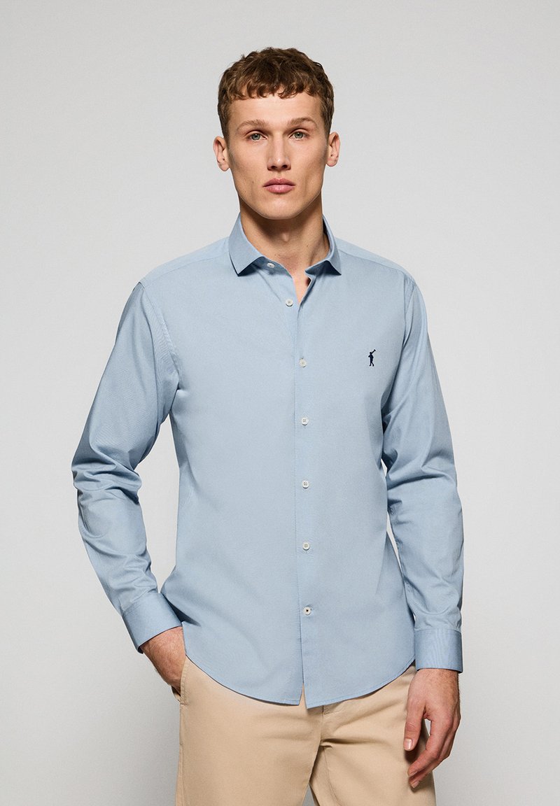 Hellblaues Langarmhemd, mit Knopfleiste, schmal geschnitten, aus Baumwollmaterial. Mit kleinem dunklem Logo auf der Brust und leichter Textur.