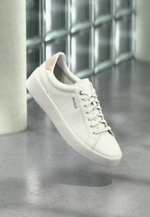 Fehér alacsony szárú sneaker fűzőkkel és bézs sarokpánttal, a "HUGO" márkajelzéssel, egy csiszolt betonpadló fölött lebeg, üvegtégla fal közelében.