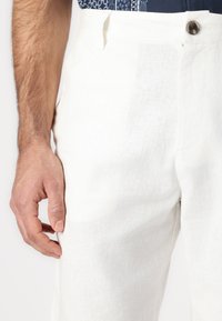 Bílé lněné kalhoty s volným střihem, zapínáním na knoflík a bočními kapsami. Tkanina má přirozenou strukturu s jemným leskem.