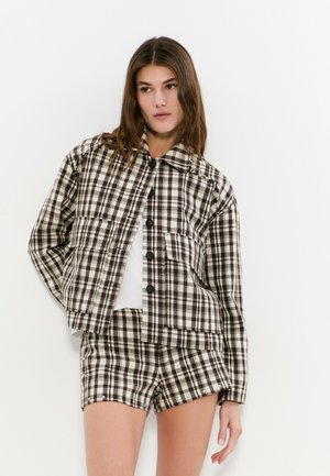 Mujer con chaqueta abotonada de cuadros marrón y blanco y pantalones cortos a juego sobre una camiseta blanca, posando frente a un fondo blanco.