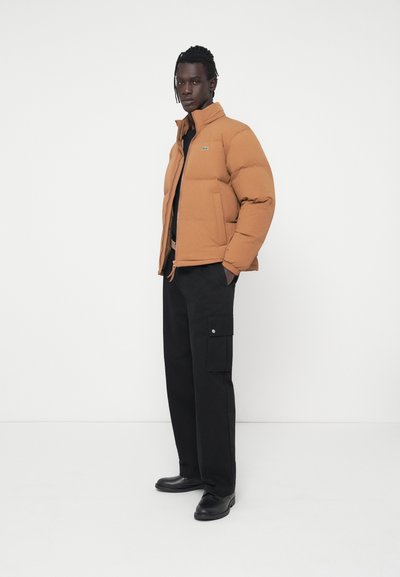 Doudoune marron avec un col haut, une fermeture éclair et des poches latérales ; associée à un pantalon cargo noir doté de poches latérales.