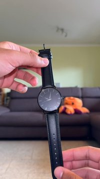 Ceas rotund negru cu un cadran gri mat, marcaje subțiri argintii pentru ore și o brățară din piele neagră cu găuri pentru ajustare.