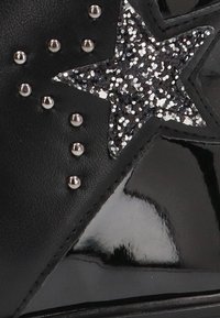 Zapato de cuero negro con un gran acento de estrella brillante, tachuelas metálicas y un acabado brillante, que resalta su superficie texturizada.