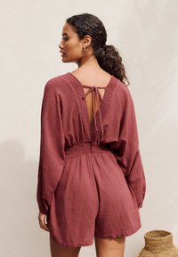 Maroon romper with long sleeves, open back design, and tie closure. Features a smocked waist and textured fabric. Complements a natural setting.