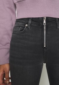 Primer plano de una persona que lleva vaqueros negros con cremallera plateada en la parte delantera y una camiseta de manga larga lavanda, con la mano relajada a un lado.