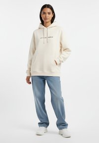 Felpa crema con testo "FACCIO IL GRANDE PASSO", tasca frontale, cappuccio con lacci, abbinata a jeans dritti azzurri chiari e sneakers bianche.