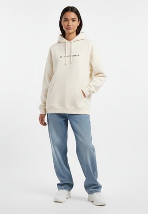 Crème hoodie met de tekst "Laten we trouwen", voorzak, capuchon met trekkoord, gecombineerd met lichtblauwe straight-leg jeans en witte sneakers.