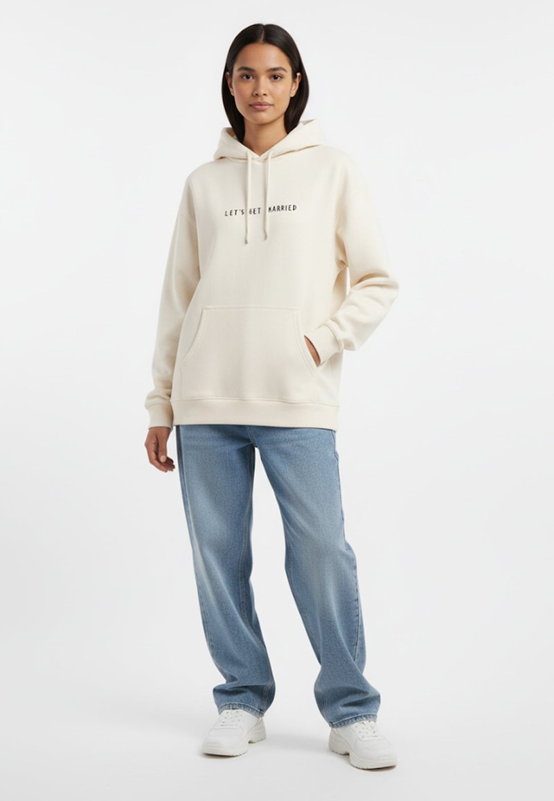 Felpa crema con testo "FACCIO IL GRANDE PASSO", tasca frontale, cappuccio con lacci, abbinata a jeans dritti azzurri chiari e sneakers bianche.