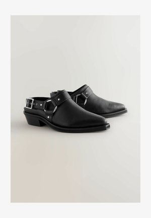 Sorte læderklipklapper med spids tå, lav hæl og justerbar rem med sølvhardware og spænde detaljer. Glat tekstur.