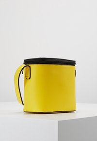 Sac circulaire jaune en matériau synthétique lisse, doté d'une fermeture éclair noire sur le dessus et d'une poignée latérale pour un transport facile.