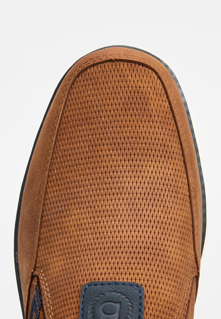 Zapato marrón sin cordones con parte superior de malla texturizada, acentos de ante suave y un parche de logo azul oscuro. La suela es oscura con un acento claro.
