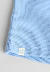 Lichtblauwe ribbelkatoenen stof met een klein crèmekleurig label met een glimlachend gezicht aan de zoom. Gladde textuur en nette stiksels zichtbaar.