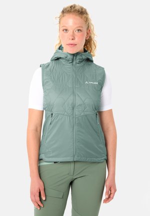 Frau trägt eine ärmellose grüne Kapuzenweste über einem weißen Kurzarmshirt und grüner Hose, steht vor einem schlichten hellen Hintergrund.