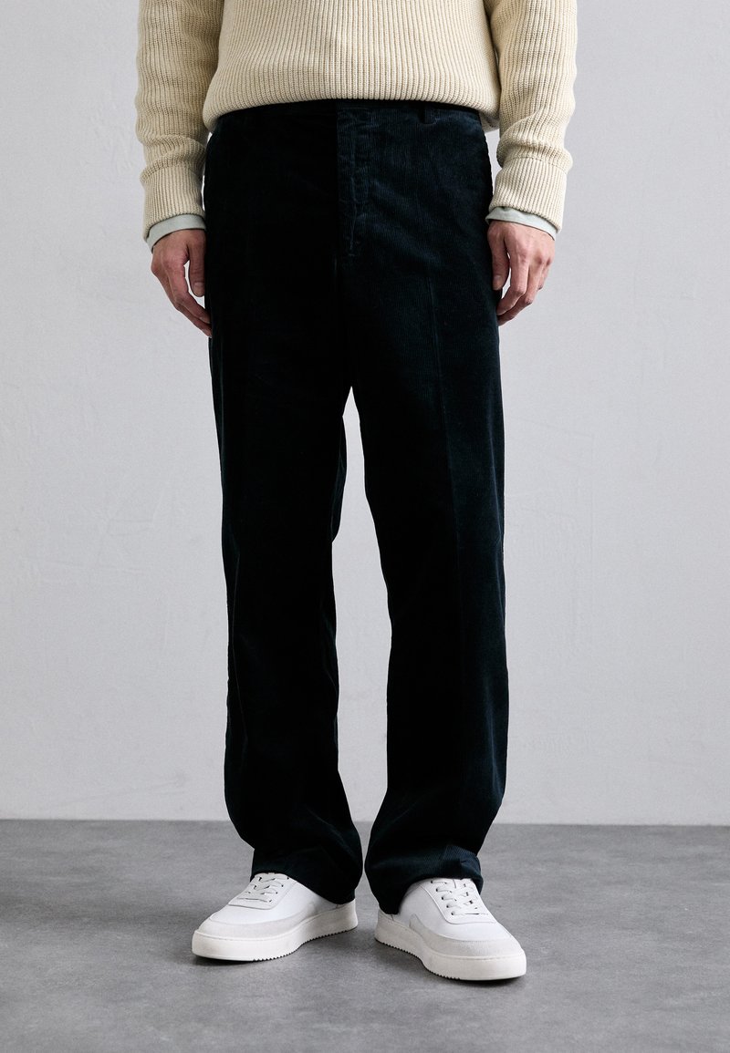 Pantalones de corduroy oscuros con un corte relajado, que presentan una superficie texturizada y un diseño de pierna recta, combinados con zapatillas deportivas blancas.