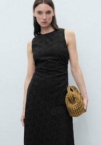 Robe sans manches noire avec broderie florale, silhouette ajustée et détail froncé sur le côté. Le mannequin tient un sac en bandoulière en tan tissé avec des poignées rondes.
