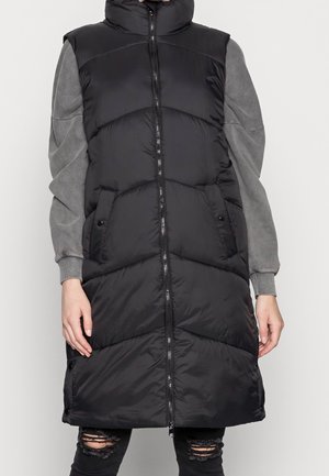 Lång svart pufferväst med hög krage, med en framdragkedja, horisontell kviltning, sidofickor och en grå tröja under.