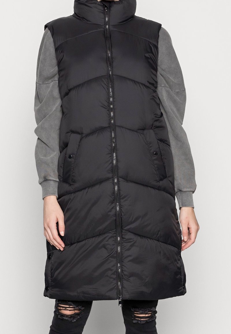 Lång svart pufferväst med hög krage, med en framdragkedja, horisontell kviltning, sidofickor och en grå tröja under.