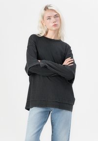 Tricou negru cu mânecă lungă, cu guler rotund, croială relaxed și margini brutale. Asortat cu blugi din denim albastru deschis, cu o textură estompată.