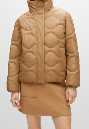 Kobieta ubrana w beżową pikowaną kurtkę typu puffer z przeszyciami falistymi i wysokim kołnierzem, zestawioną z dopasowaną beżową wełnianą mini spódnicą.