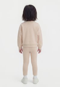 Otrok s ravnimi temnimi lasmi stoji obrnjen stran, oblečen v bež pulover in hlače v ujemajoči se barvi, z belimi nogavicami in supergami na belem ozadju.