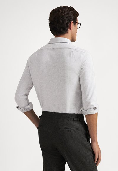 Άνδρας με σκούρα μαλλιά και γυαλιά, που φοράει ανοιχτό γκρι μακρυμάνικο πουκάμισο με ανασηκωμένα μανίκια και σκούρο γκρι παντελόνι, στέκεται μπροστά σε λευκό φόντο.