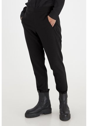 Personne portant un pantalon noir slim avec des poches latérales et des bottines noires épaisses, debout sur un fond clair uni.