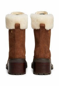 Botas de tobillo de ante marrón con un cuello de piel sintética beige, con un tacón ancho y un detalle del logo dorado en la parte trasera. Acentos texturizados en toda la bota.