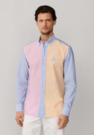 Hombre de pie mirando hacia adelante, vistiendo una camisa de manga larga con botones y bloques de color en tonos pastel rosa, azul y durazno, y pantalones blancos.