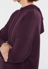 Lila gestreifte Kapuzenjacke mit gerippter Oberfläche, ausgestattet mit einer Kordelzugkapuze und lockerer Passform. Der Stoff wirkt weich und leicht dehnbar.