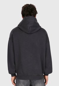 Person med krøllet mørkt hår iført en ensfarvet sort hoodie og slidte sorte jeans, set fra ryggen mod en ensfarvet baggrund.