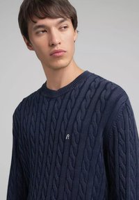 Marineblauwe kabelgebreide trui met ronde hals en geribbelde manchetten. Klein logodetail op de borst, met een textuurpatroon over het hele kledingstuk.