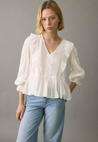 Bluză albă peplum cu detalii din volane pe umeri, decolteu în V și închidere cu nasturi; asortată cu blugi din denim bleu deschis.