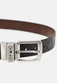 Schwarzer synthetischer Gürtel mit geprägtem Muster, silberner Metallschnalle mit dem Wort "GUESS" und braunem Innenfutter.