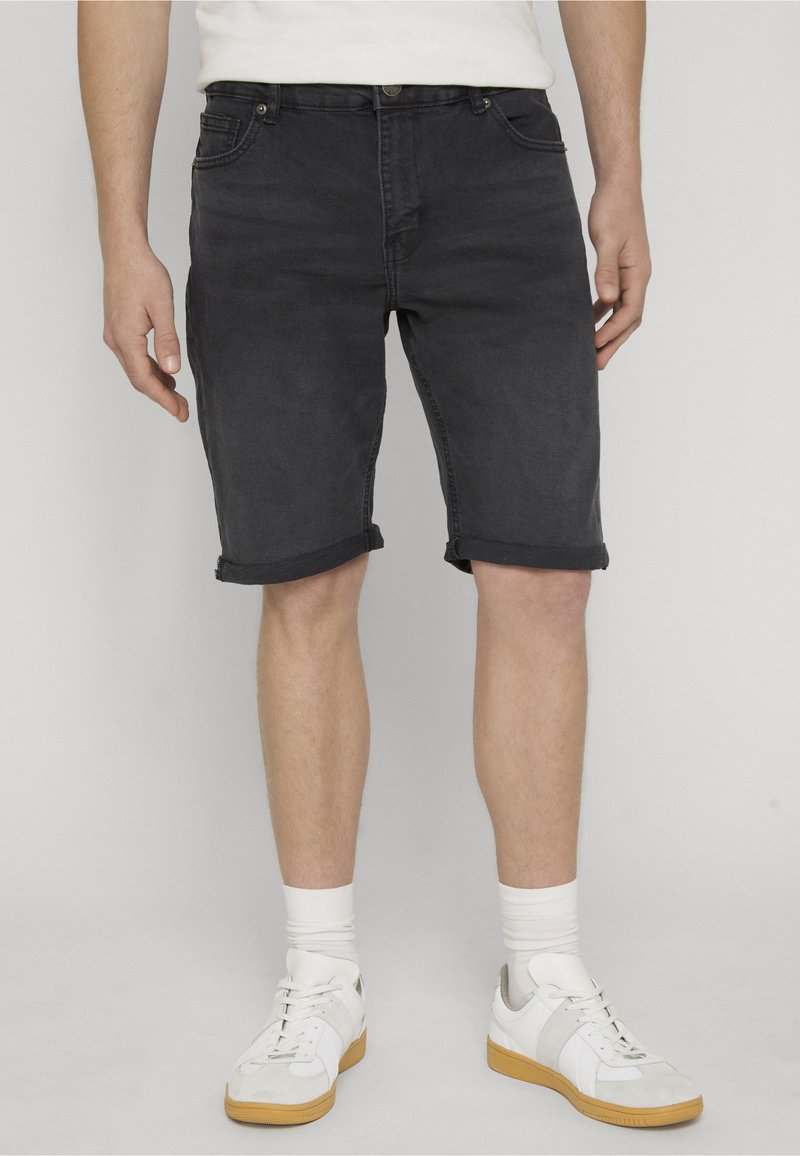 Homme portant un short en jean noir retroussé, des chaussettes blanches et des baskets blanches, debout devant un fond uni.