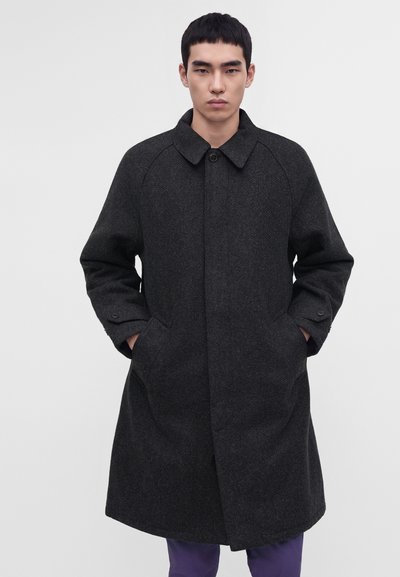 Young man standing with hands in pockets wearing a dark gray long wool coat with a collar and button closure.