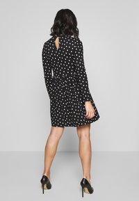Czarna sukienka w białe kropki z długim rękawem, marszczonym pasem, detalem w kształcie dziurki z tyłu oraz rozkloszowaną spódnicą; połączona z czarnymi szpilkami.