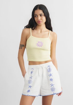 Hosszú sötét hajú nő, sárga "Santa Cruz" logós crop topot és fehér rövidnadrágot visel kék tengeri állatmintával, kezét zsebre dugva.