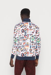 Färgglad bomberjacka med ett vitt tyg dekorerat med olika logotyper och lappar. Ribbad mudd och krage i marinblått.