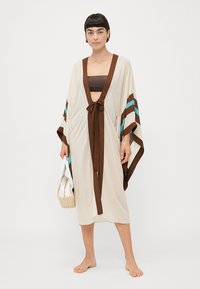 Robe de style kimono en crème, avec des rayures marron et teal. Présente une fermeture à nouer, des manches larges et un tissu léger et fluide.