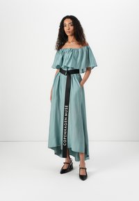 Abito turchese senza spalle con maniche a campana, lungo orlo asimmetrico, cintura nera con la scritta "COPENHAGEN MUSE" e scarpe col tacco nere.