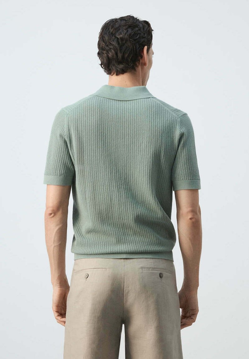 Homme de dos portant un polo en maille vert clair à manches courtes et un pantalon beige sur un fond uni clair.