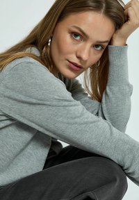 Maglione grigio a maniche lunghe con polsini a costine, indossato da un modello con capelli lunghi. Tessuto texturizzato, abbinato a pantaloni neri, per un look rilassato.