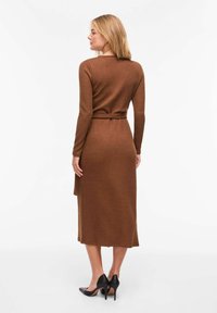 Robe portefeuille marron à manches longues avec ceinture à nouer, longueur au genou, en matériau doux. La tenue est complétée par des chaussures noires à talons hauts.