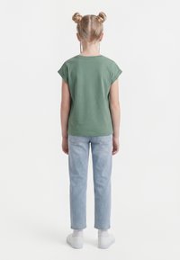 Niño con cabello rubio recogido en dos moños, vestido con una camiseta verde de manga corta, jeans azul claro, calcetines blancos y zapatillas blancas, visto de espaldas.