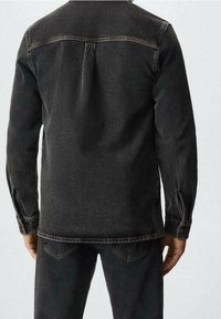 Chaqueta de mezclilla negra con corte recto, detalles de costura visibles y puños abotonados. La textura está ligeramente desgastada, presentando un clásico yugo trasero.